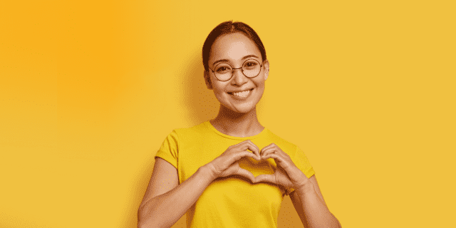 Femme portant un chandail jaune sur fond jaune faisant un coeur avec ses main au niveau de la poitrine